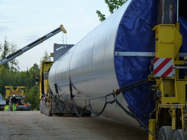Bild vergr&ouml;&szlig;ern: Schwertransport eines großen Windkraft‑Turmsegments auf einem Spezialanhänger. Das silberne Bauteil mit blauer Schutzabdeckung wird auf einem gelben Transportgestell gesichert und von Kränen sowie Begleitfahrzeugen umgeben. Arbeiter in Warnkleidung koordinieren den Transport auf einem Industrie‑ bzw. Baustellengelände. Die Szene zeigt die professionelle Logistik beim Transport übergroßer Komponenten für Windenergieanlagen.